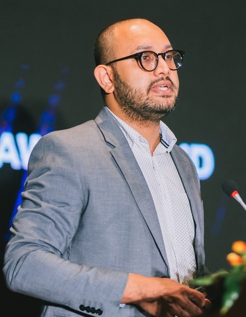 Lesith Dissanayaka speaking on stage at an event, wearing glasses, a grey jacket over a white shirt, and in front of a back screen.